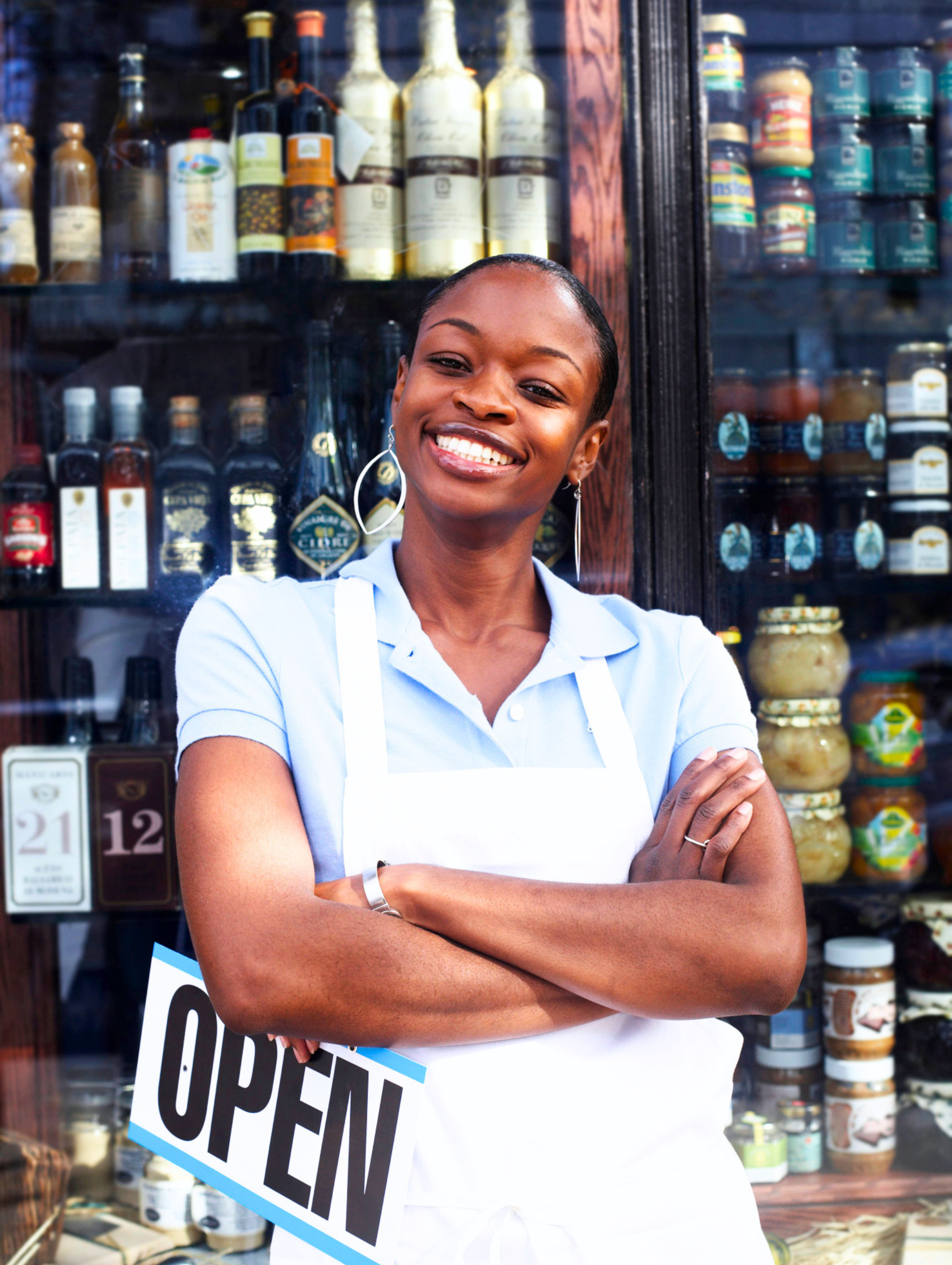 A smiling person in a white apron stands with arms crossed in front of a shop window, holding an