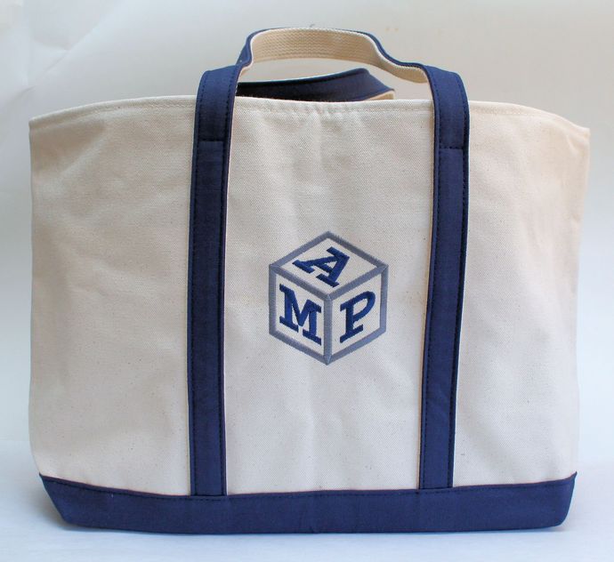 A cream-colored canvas tote bag with navy blue straps, base, and an embroidered cube monogram featuring the letters AMP. A cream-colored canvas tote bag with navy blue straps, base, and an embroidered cube monogram featuring the letters AMP.