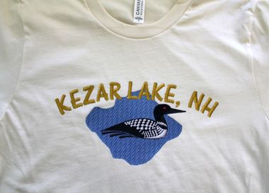 A cream-colored t-shirt with an embroidered blue lake shape, a black and white loon, and the text A cream-colored t-shirt with an embroidered blue lake shape, a black and white loon, and the text