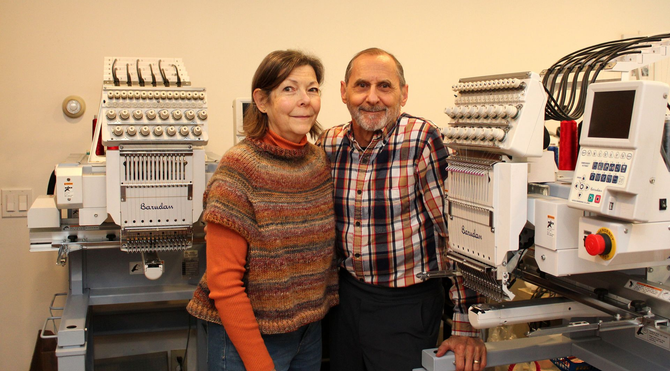 A man and a woman stand smiling between two industrial embroidery machines in a room with light-colored walls. A man and a woman stand smiling between two industrial embroidery machines in a room with light-colored walls.