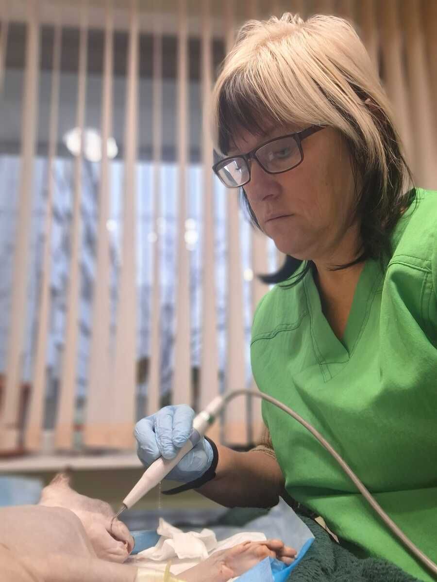 Zobārstniecības klīnicists zaļā darba apģērbā, izmantojot rokas instrumentu procedūras laikā, valkājot cimdus un brilles.
