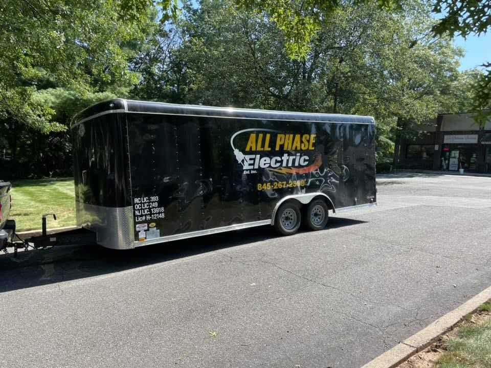 Black enclosed trailer with “ALL PHASE Electric” logo parked on a tree-lined road.
