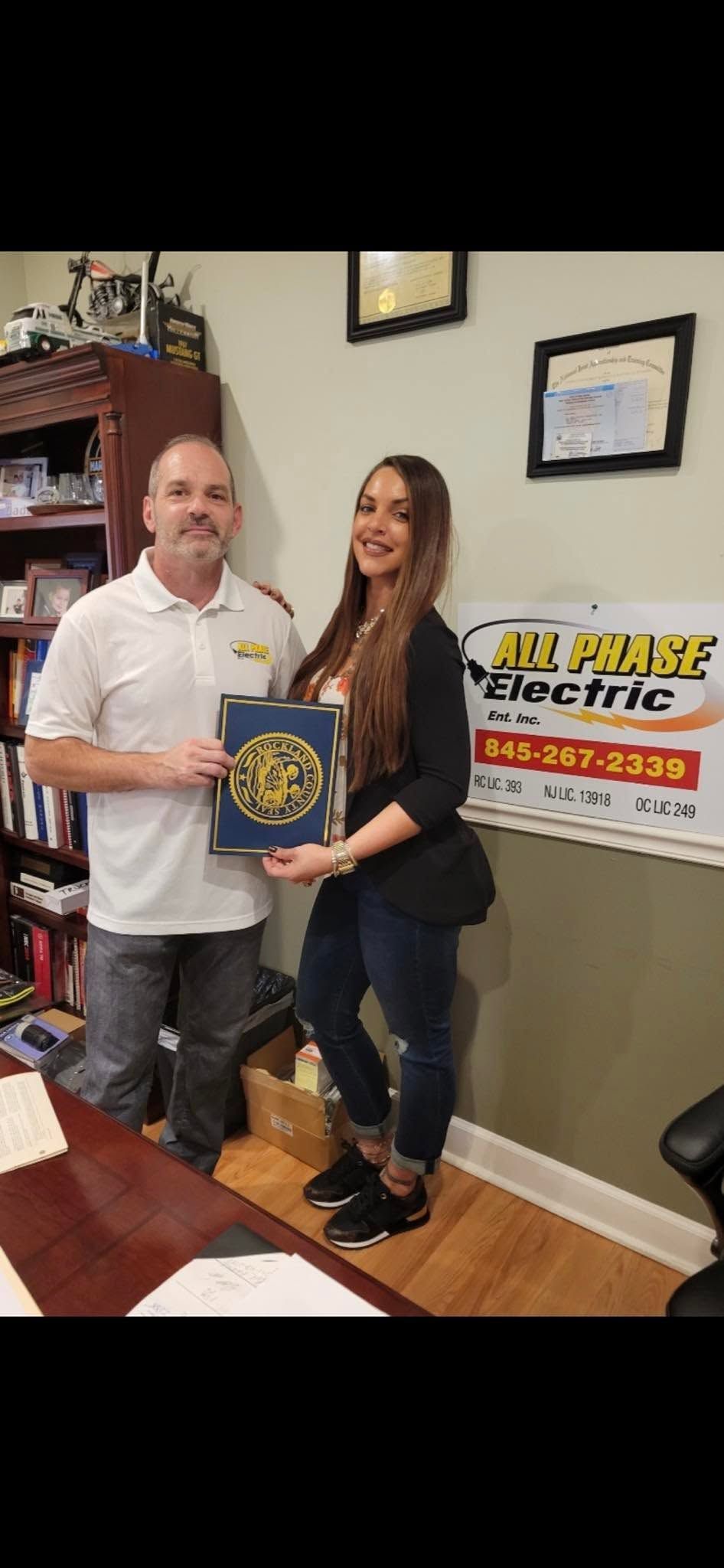 Two people pose indoors holding an award plaque beside a wall sign for a local business.