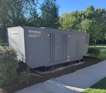 Outdoor generator beside a house deck on a grassy yard, with a vented beige cover and white base.