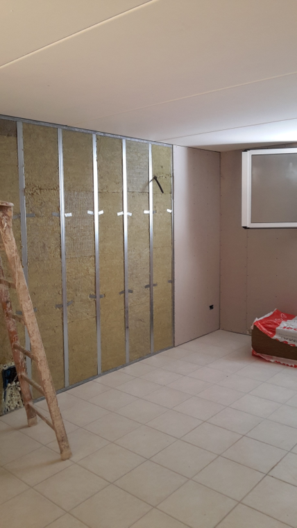 Una scala di legno si erge davanti a un muro in costruzione con montanti metallici e pannelli isolanti fonoassorbenti gialli.