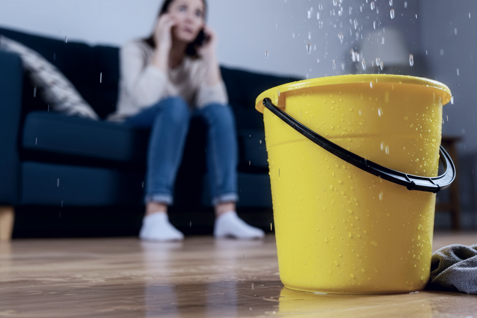 Water leaking into a yellow bucket on a hardwood floor; woman on the couch, distressed on the phone.