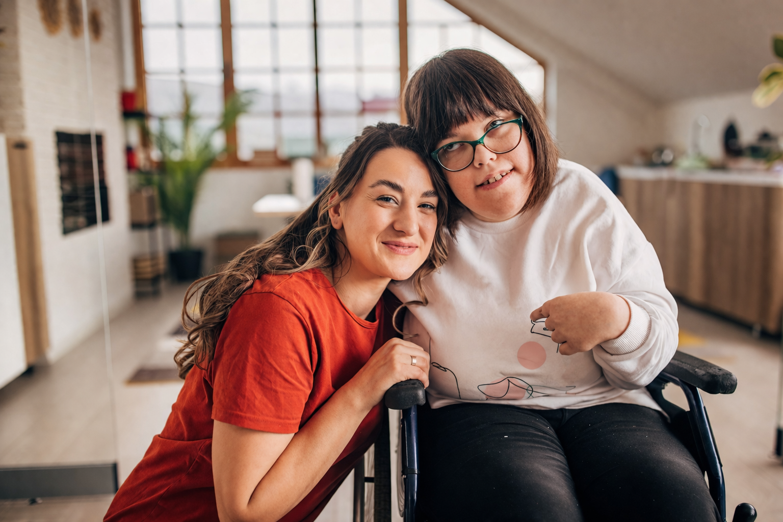 Two people sitting together indoors, smiling and leaning close in a bright living room, one is in a wheelchair