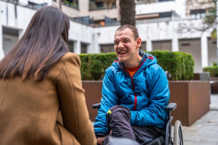 Two people talking outdoors, one seated in a wheelchair, wearing a blue jacket and smiling