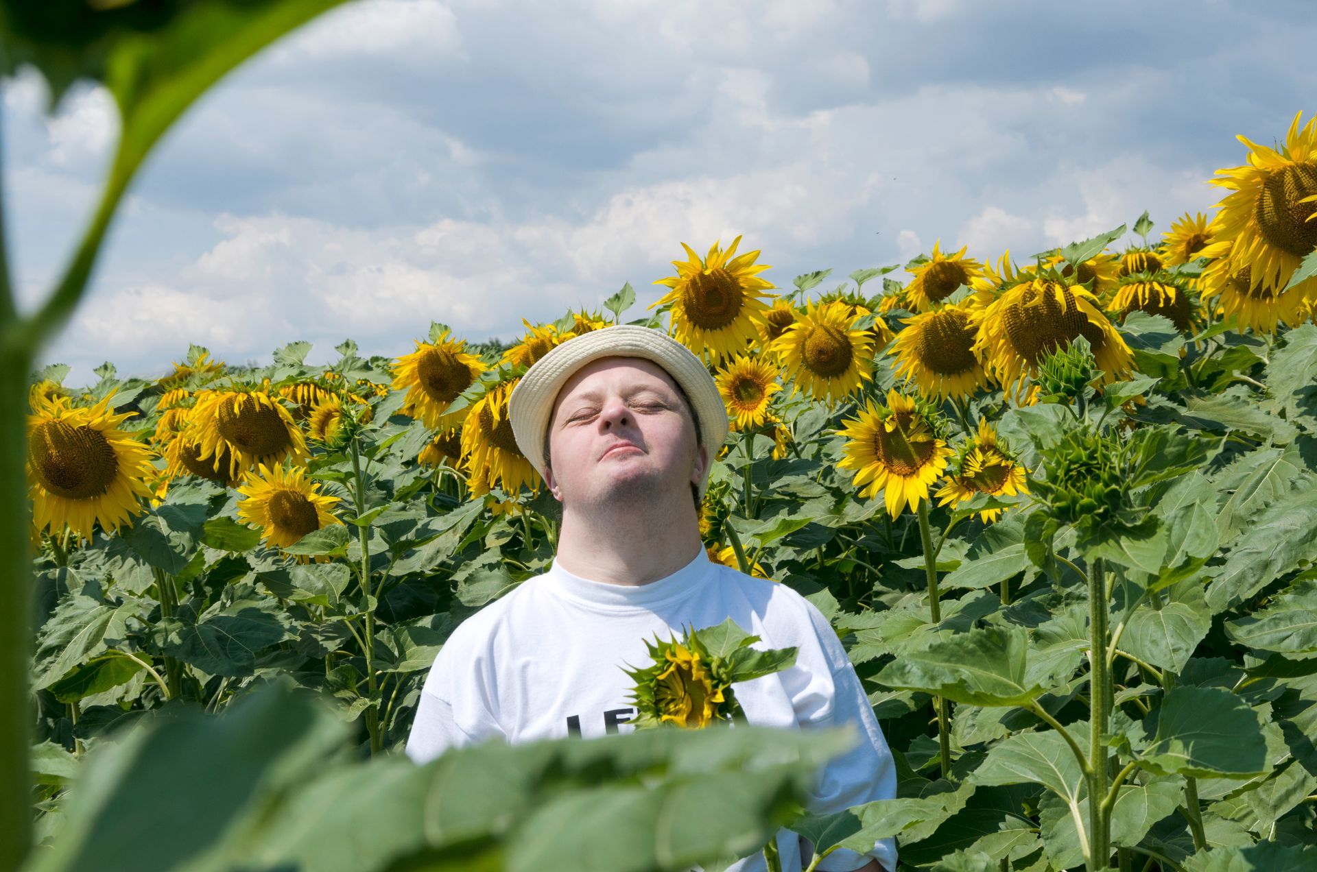 Person with a disability standing in a sunflower field with eyes closed, holding a sunflower under a cloudy sky