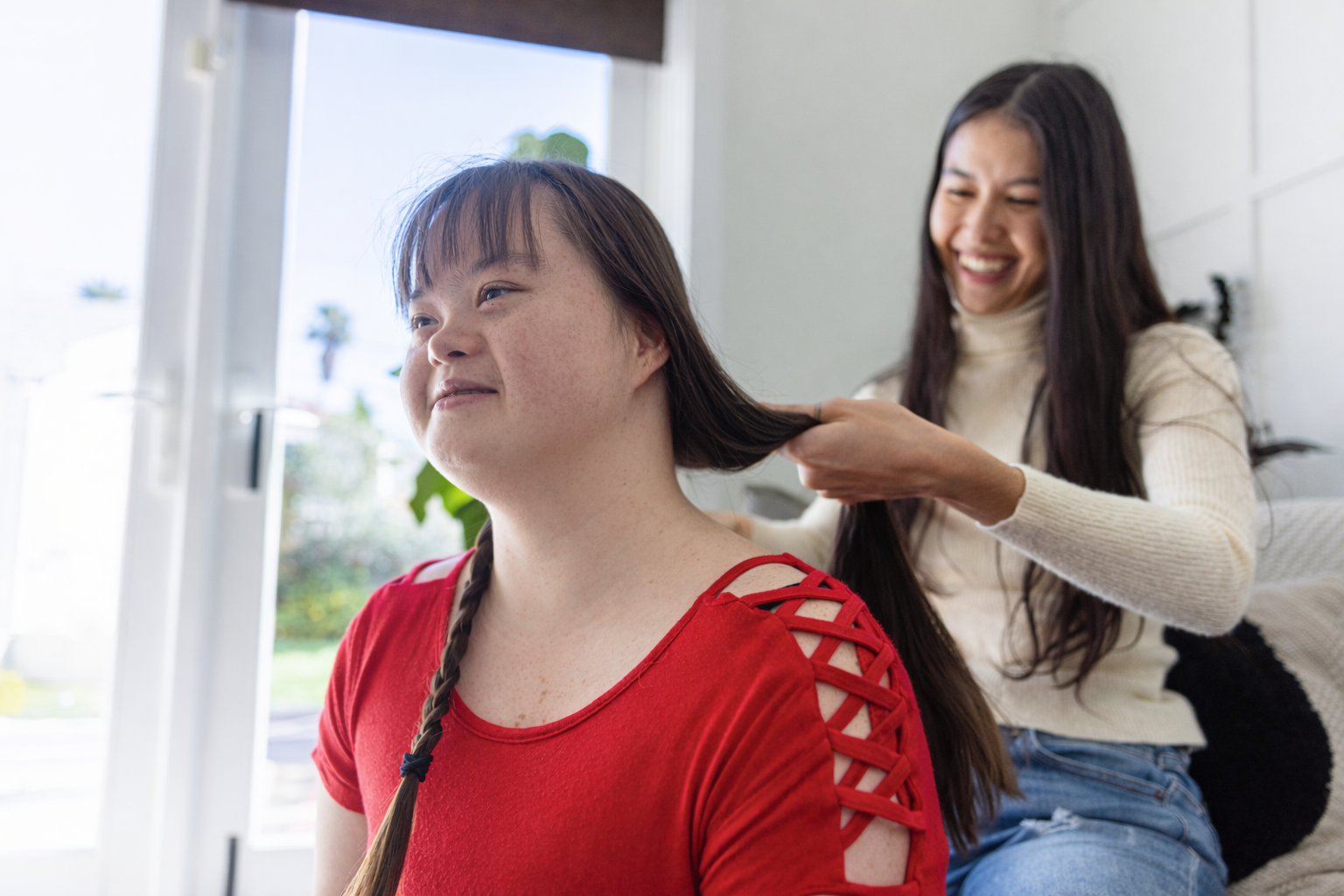 A woman braids another woman's hair, who has a disability, in a bright room.