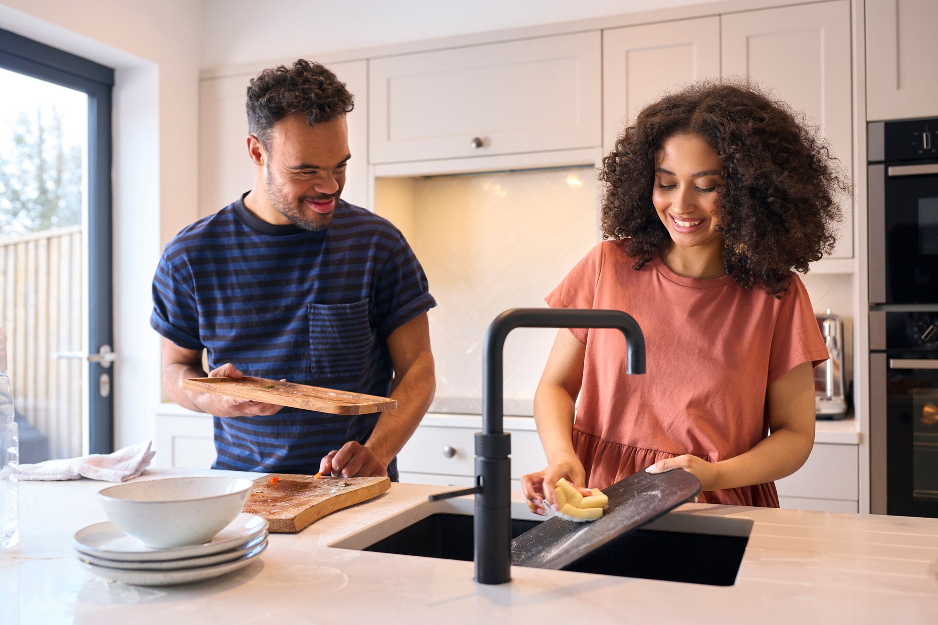 A support worker and a person with a disability are preparing food in a bright kitchen, slicing bread at a counter with bowls nearby.