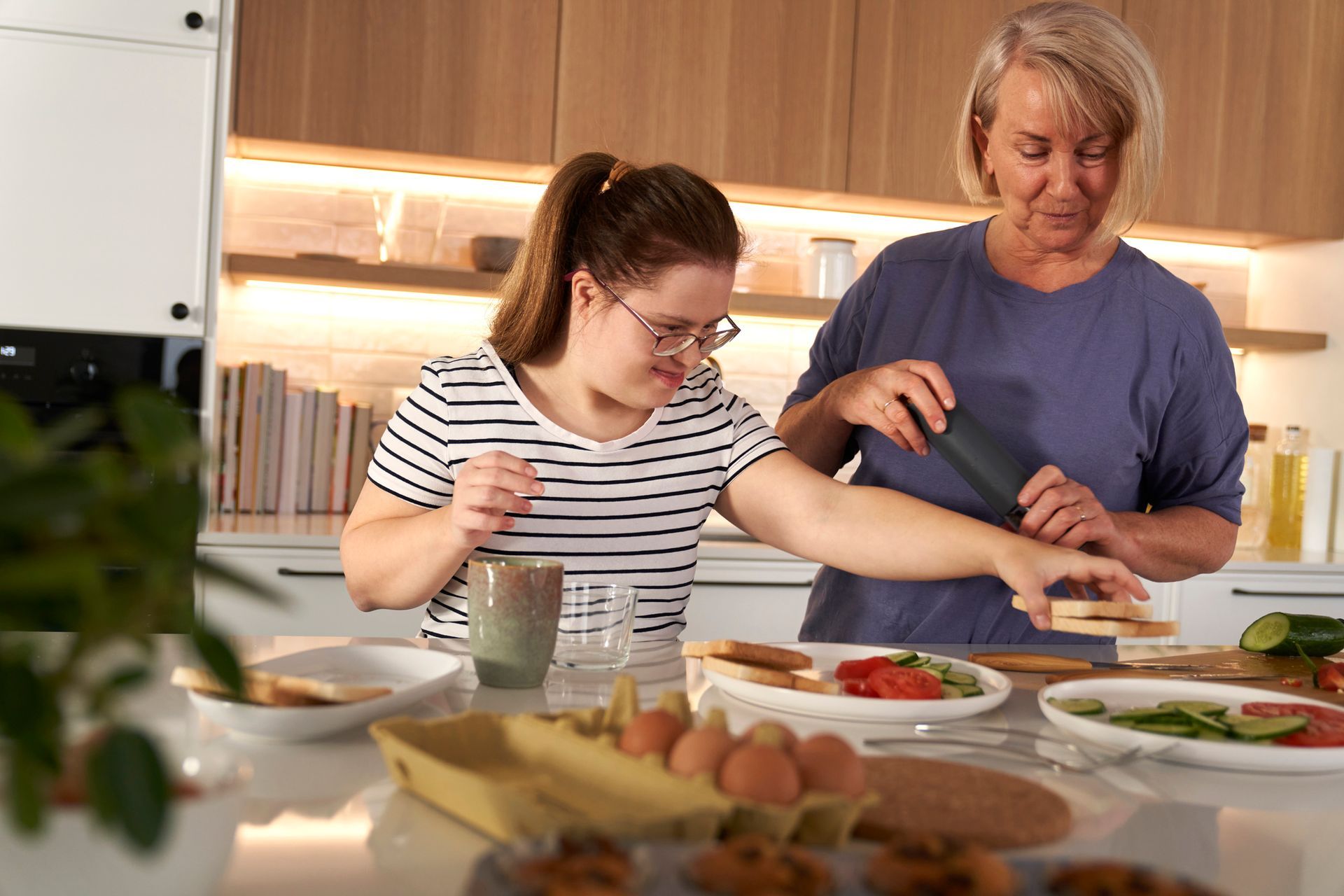 A support worker and a person with a disability are preparing food together at a kitchen counter, with plates and ingredients spread out