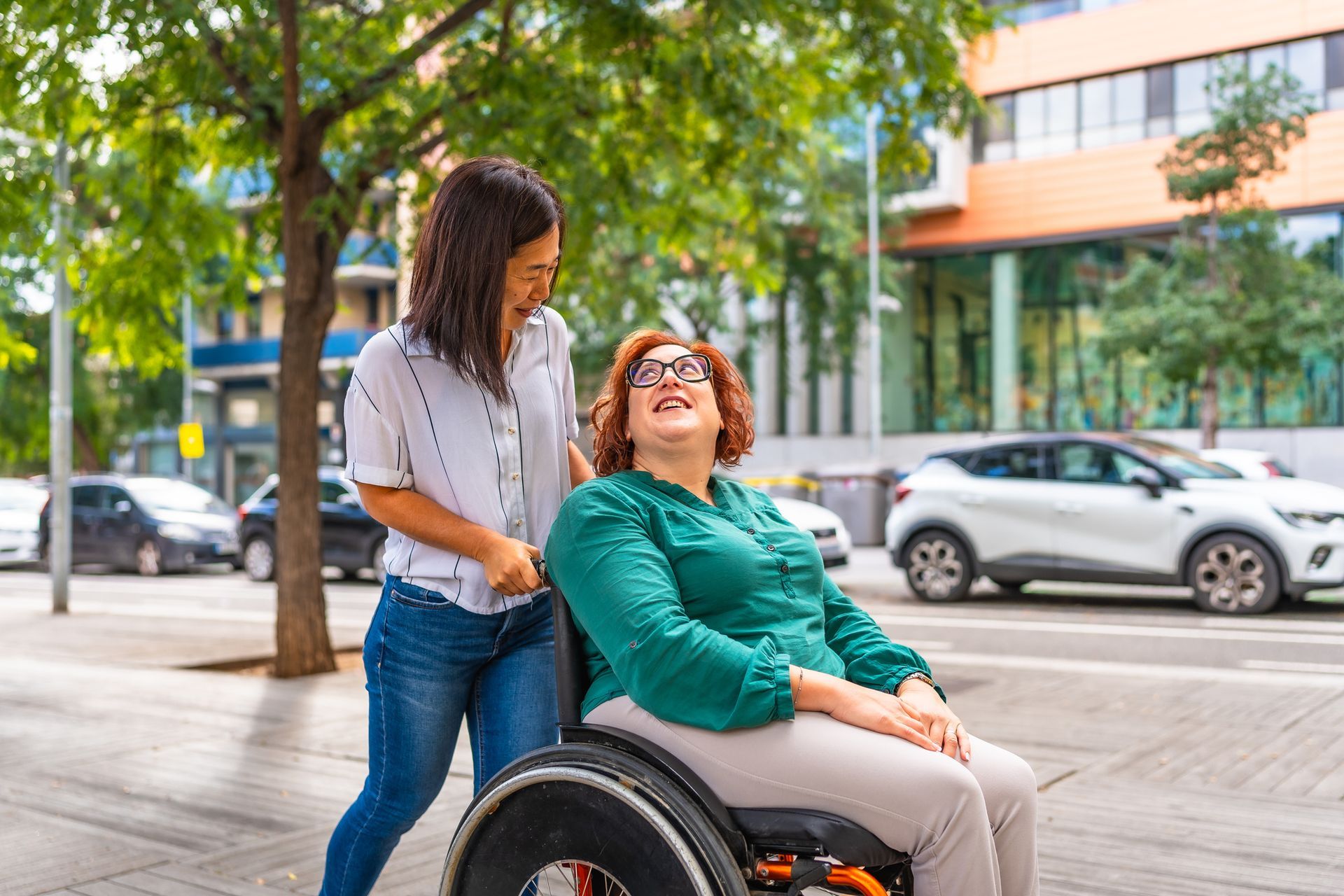 Woman assisting a wheelchair user on a tree-lined city sidewalk near parked cars