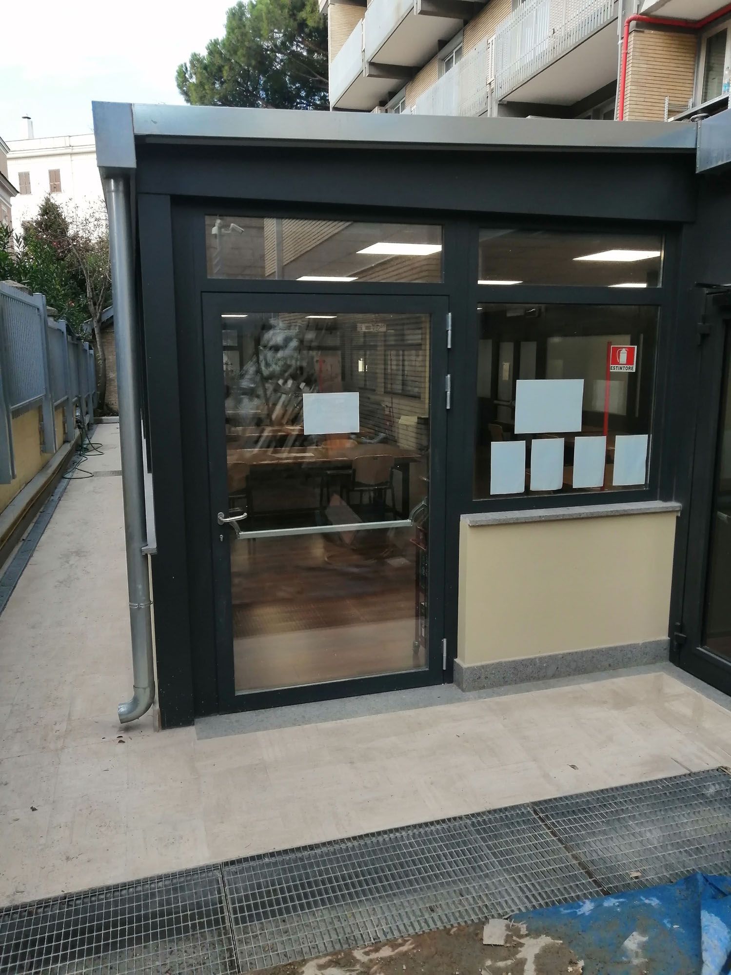Vista esterna di un edificio con porte e finestre in vetro, telaio nero. Muro beige, sentiero in cemento.