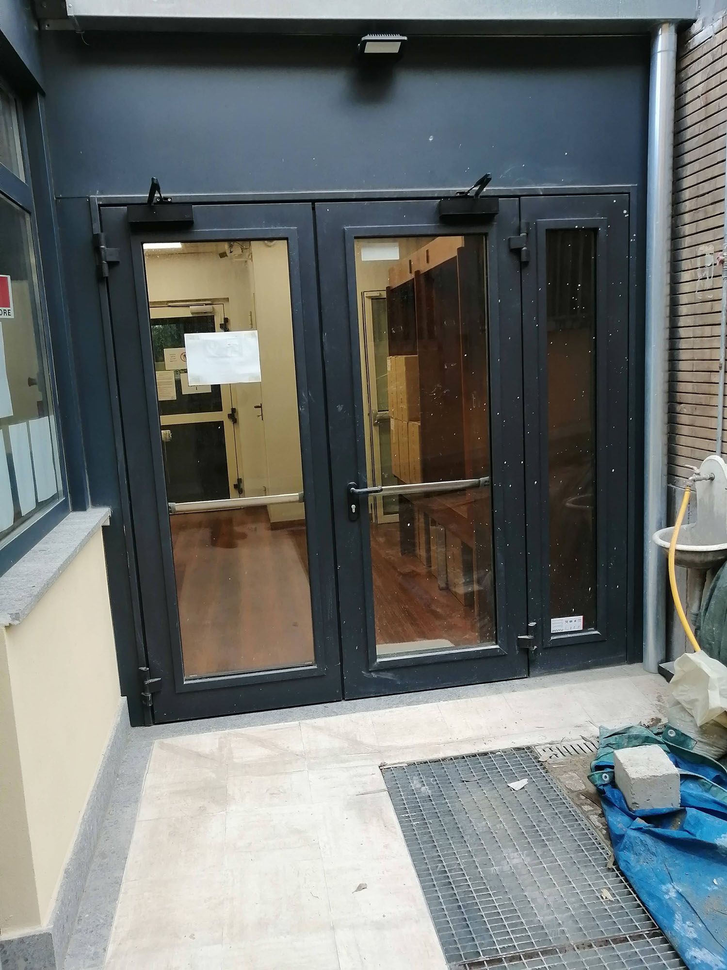Porte in vetro nero con maniglie a leva, chiusure automatiche e un piccolo zerbino su un patio piastrellato.