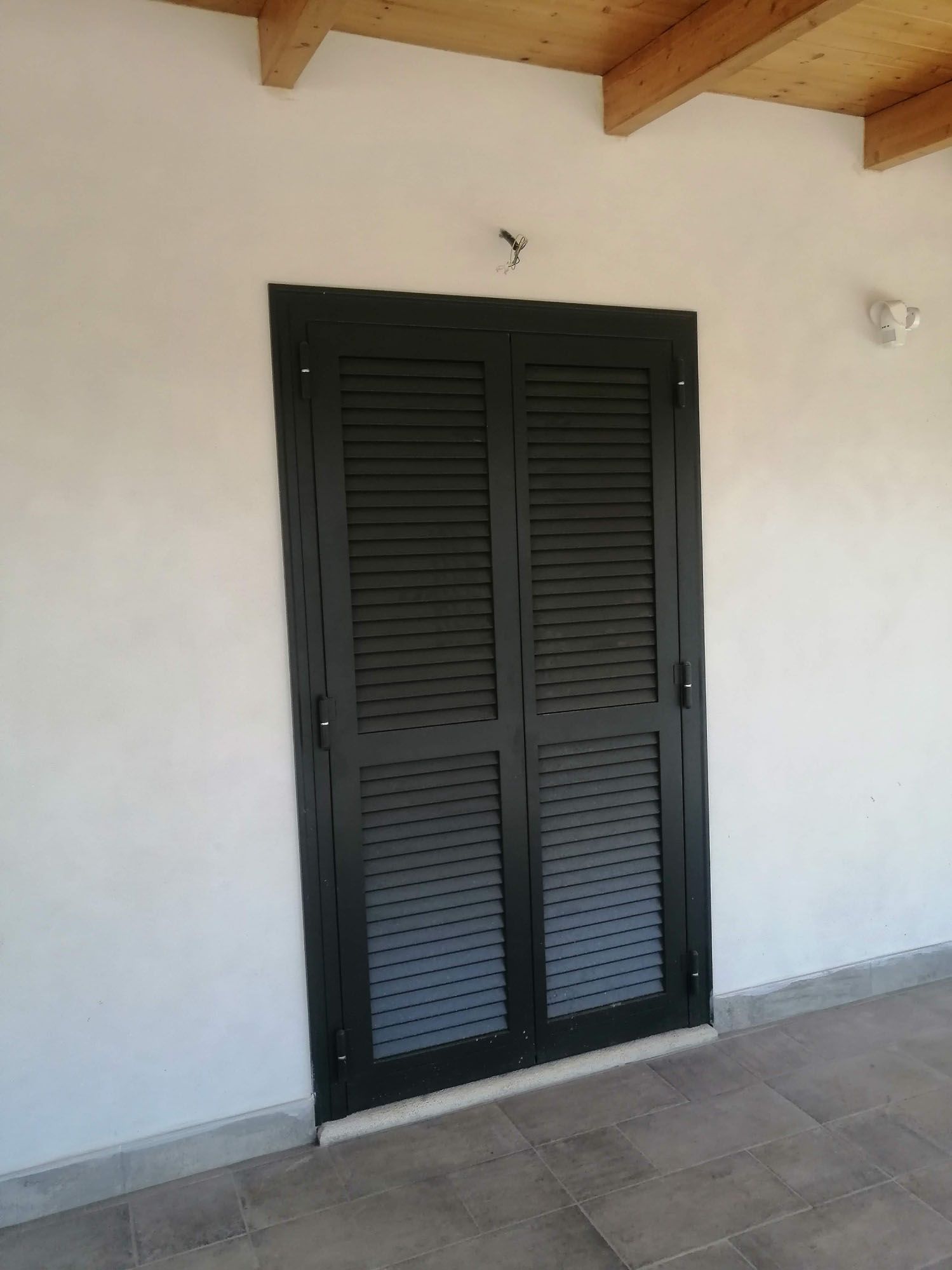 Porta con persiane nere su una parete bianca sotto una sporgenza in legno.