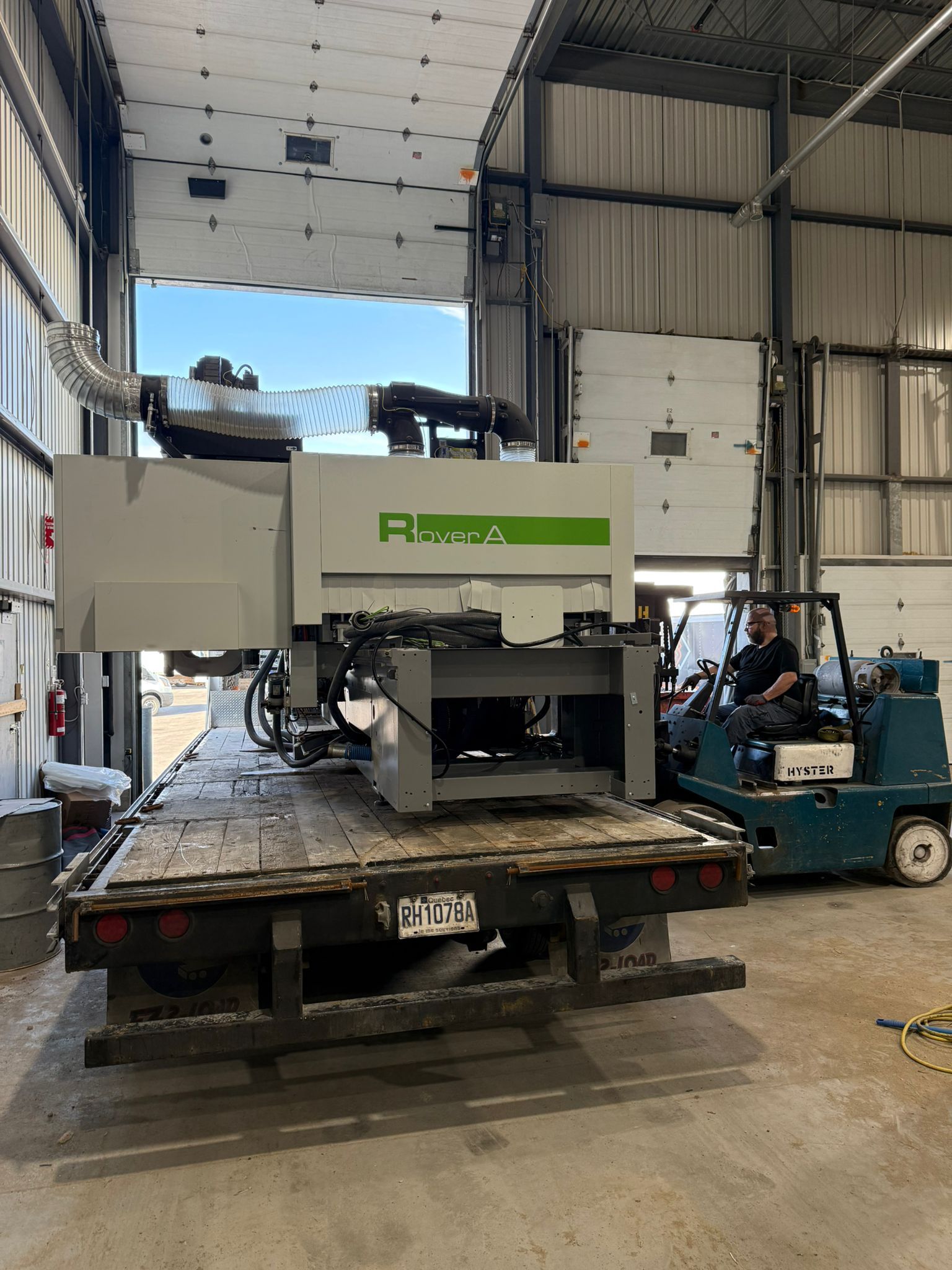 Une grande machine industrielle sur un camion à plateau est déchargée avec un chariot élévateur à l'intérieur d'un entrepôt.