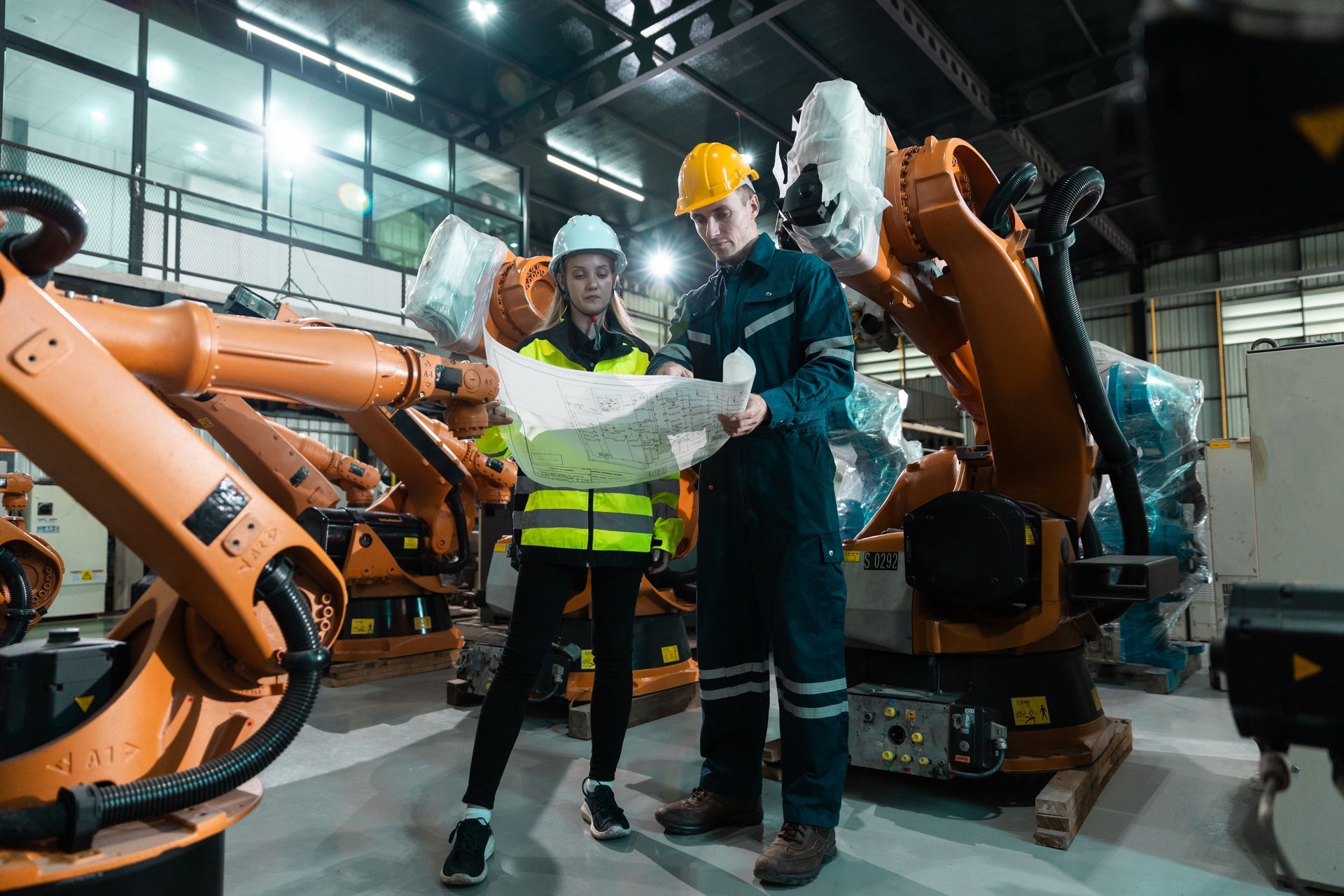Deux ingénieurs portant des casques de chantier examinent des plans dans une usine équipée de bras robotisés.