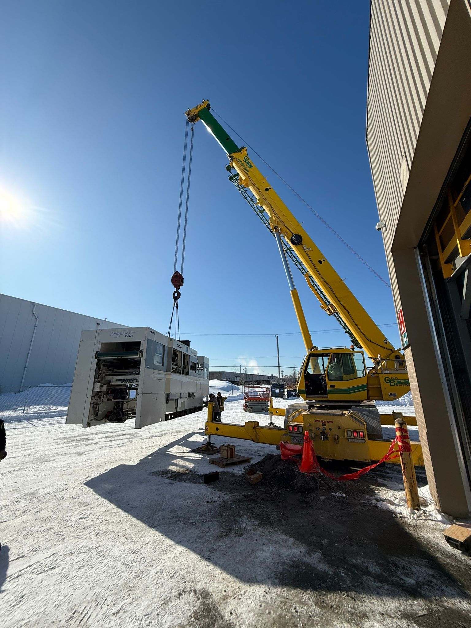 Grue jaune soulevant de grosses machines à l'extérieur d'un bâtiment par une journée enneigée.