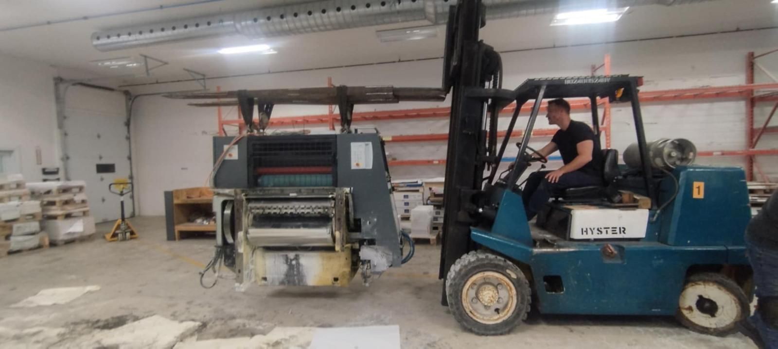 Un homme dans un chariot élévateur transporte des machines lourdes à l'intérieur d'un entrepôt.