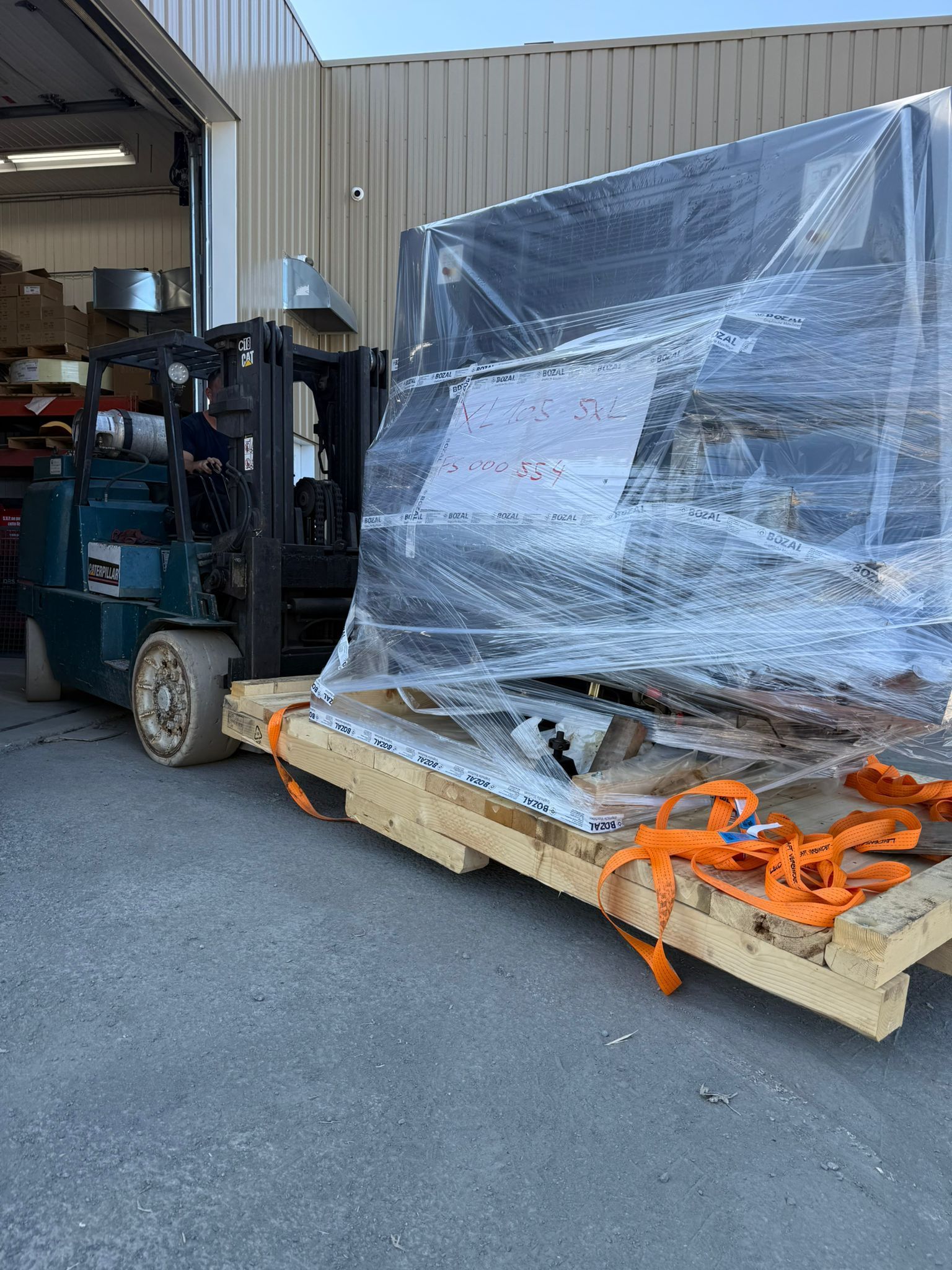 Chariot élévateur transportant un objet volumineux emballé sous plastique sur une palette en bois à l'extérieur d'un entrepôt. Des sangles orange maintiennent la charge.