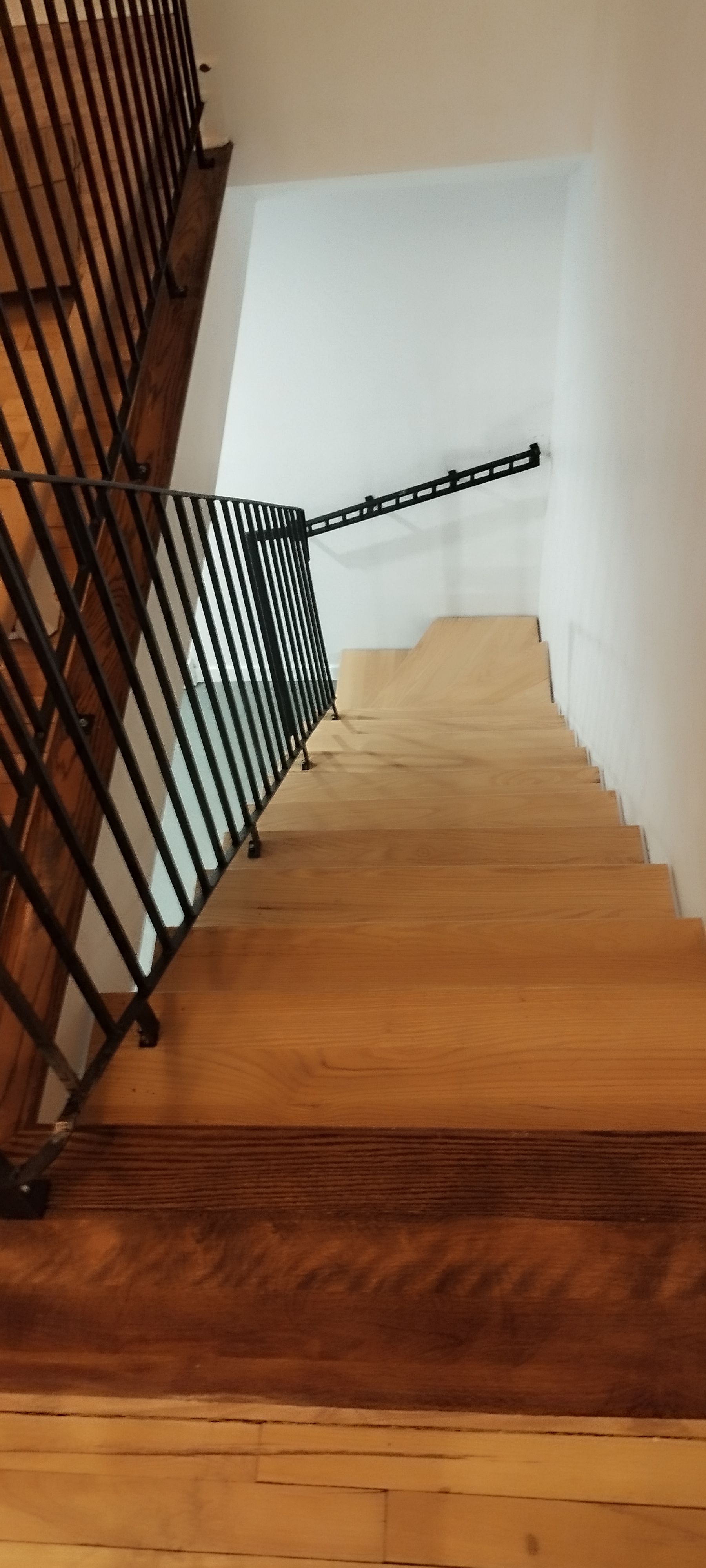 Escalier en bois avec rampe en métal noir, descendant. Murs blancs.
