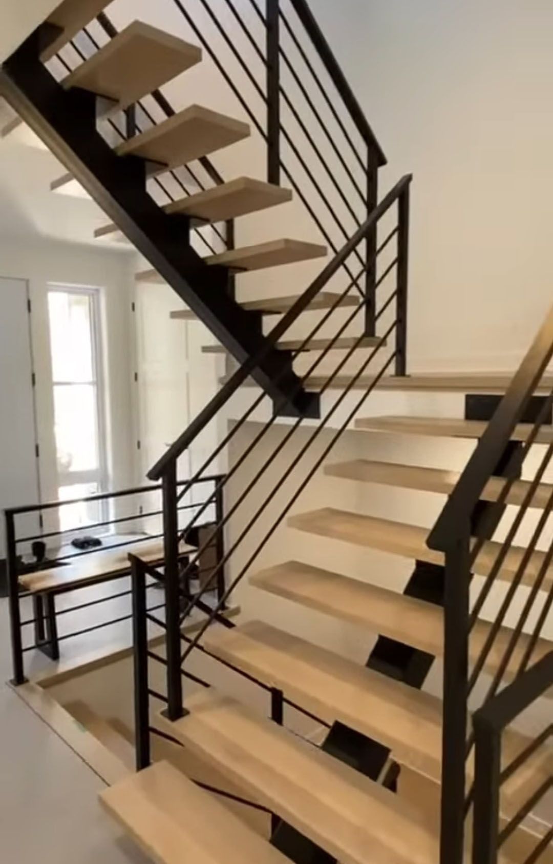 Escalier moderne avec marches en bois clair, rampes en métal noir, dans une pièce blanche lumineuse.
