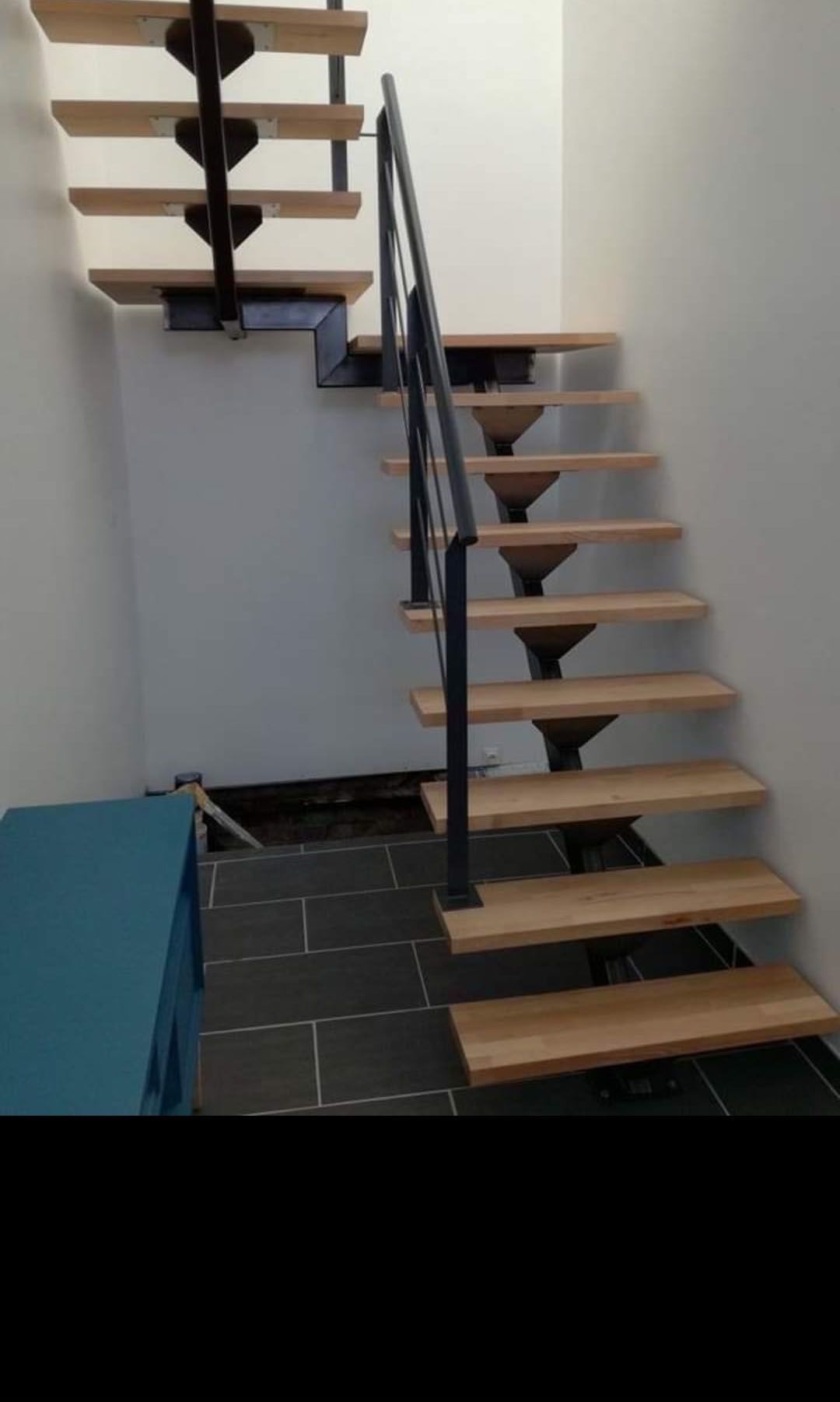 Escalier en bois avec supports et rampe en métal noir. Sol carrelé gris, armoire bleue en dessous.