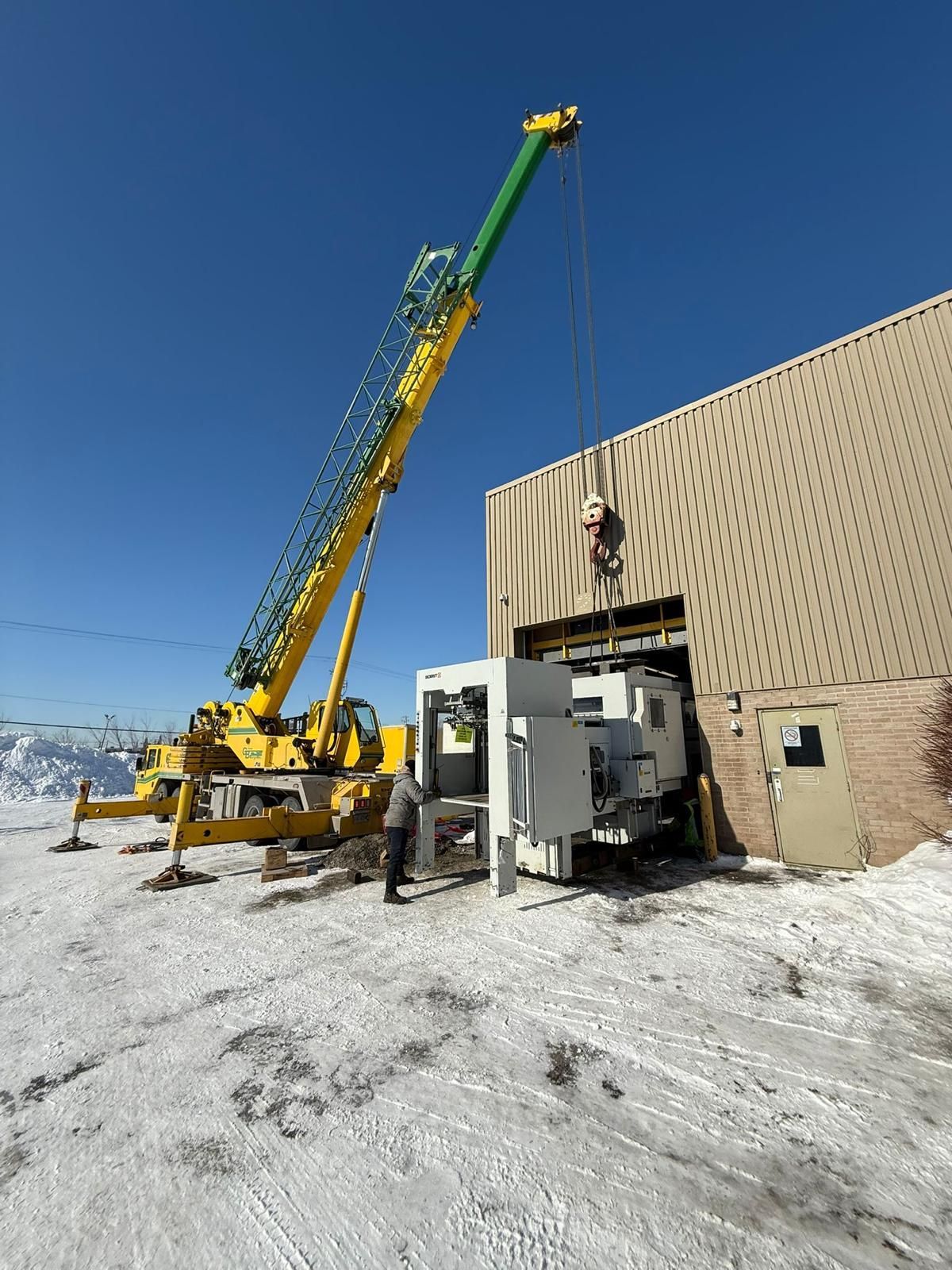 Une grue jaune soulève du matériel dans un bâtiment avec une porte de baie partiellement ouverte ; neige au sol.