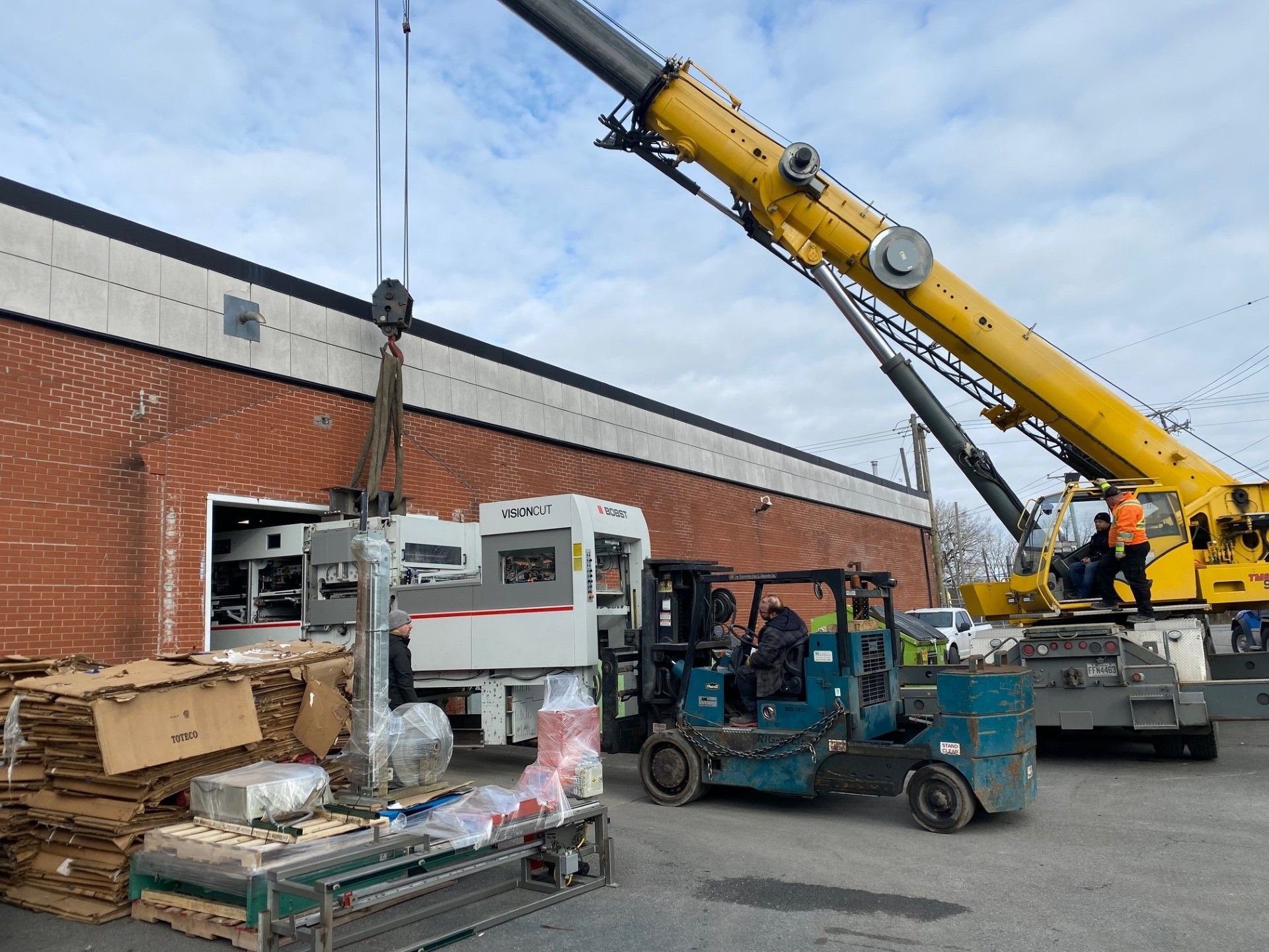 Une grue soulève des engins à l'extérieur d'un bâtiment en briques ; un chariot élévateur et des cartons sont présents.