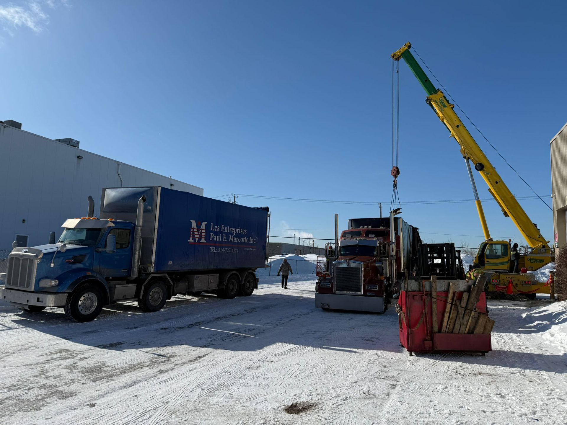Deux camions et une grue dans une zone enneigée, avec une personne marchant à proximité. Le ciel est bleu.