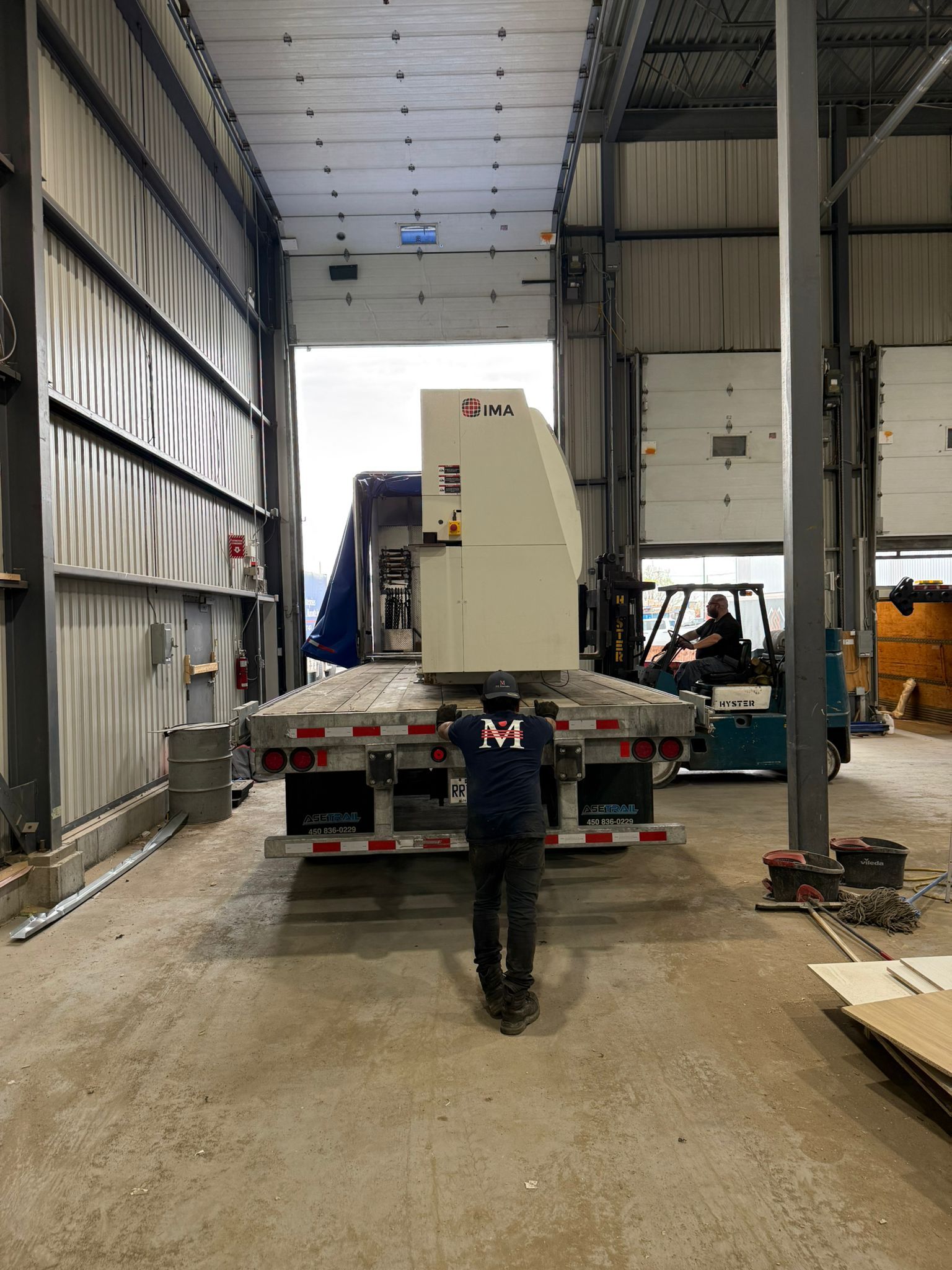 Un homme guide des machines hors d'un camion sur un quai de chargement ; chariot élévateur en arrière-plan.