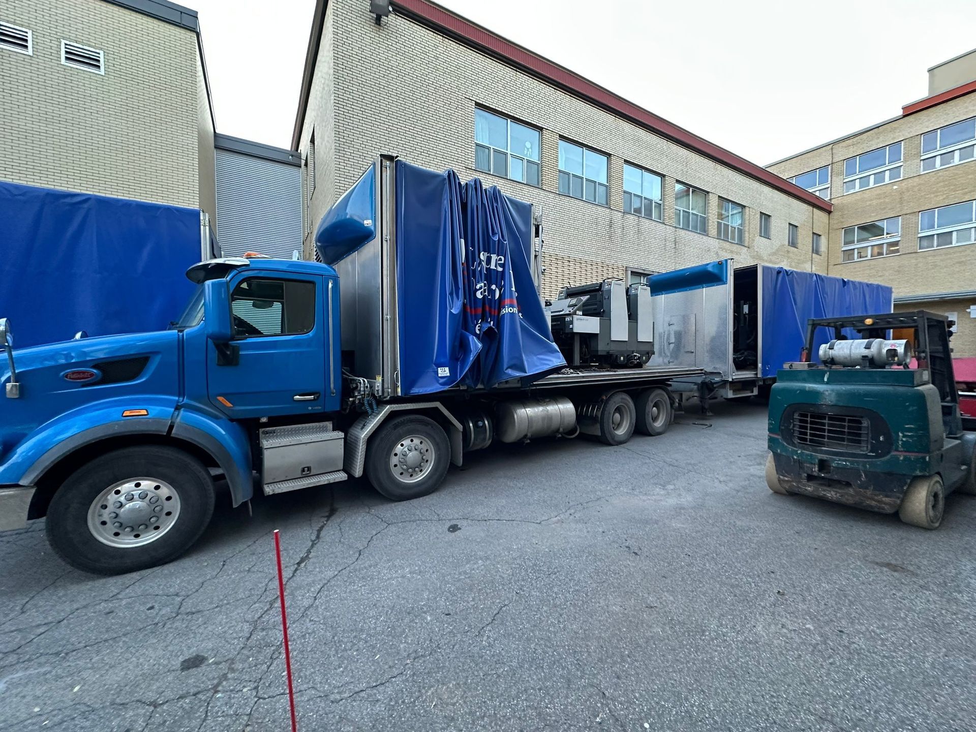 Semi-remorque bleu avec chargement, garé à côté d'un bâtiment. Un chariot élévateur est stationné à proximité.