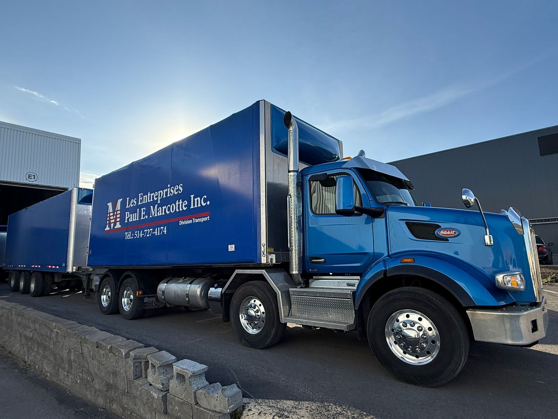 Semi-camion bleu avec remorque attachée, stationné près d'un quai de chargement; soleil éclatant.