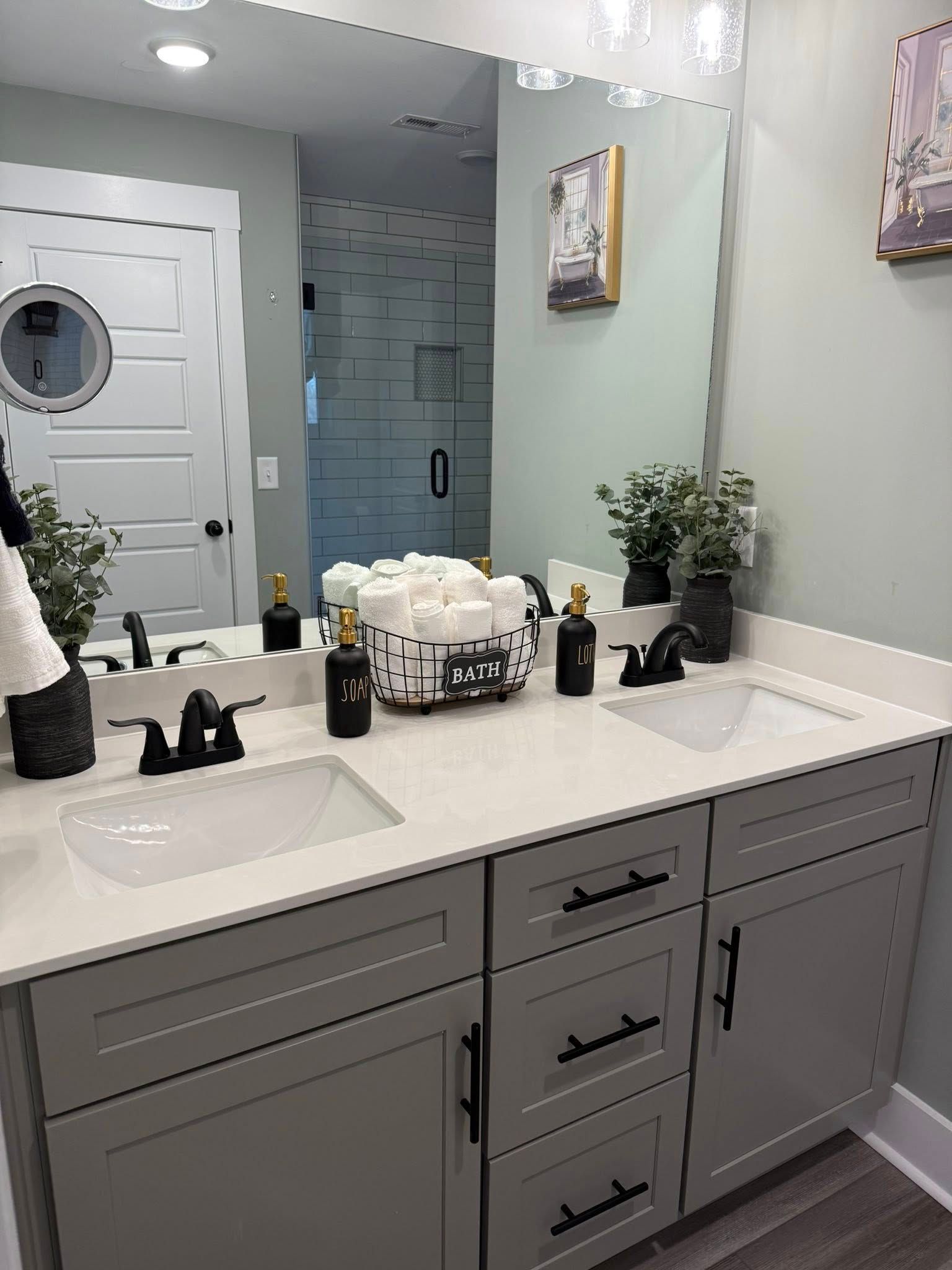 Bright bathroom vanity with double sinks, gray cabinets, mirror, decor, and a door reflected in the mirror.