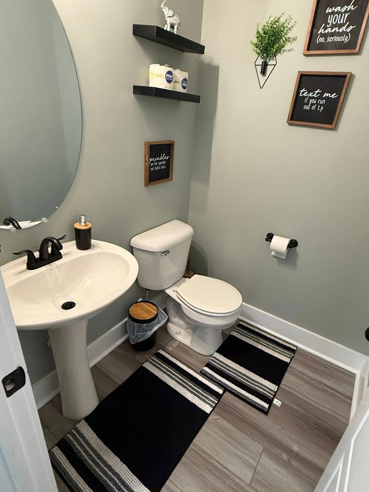 Small gray bathroom with pedestal sink, toilet, striped rug, wall shelves, and framed signs.