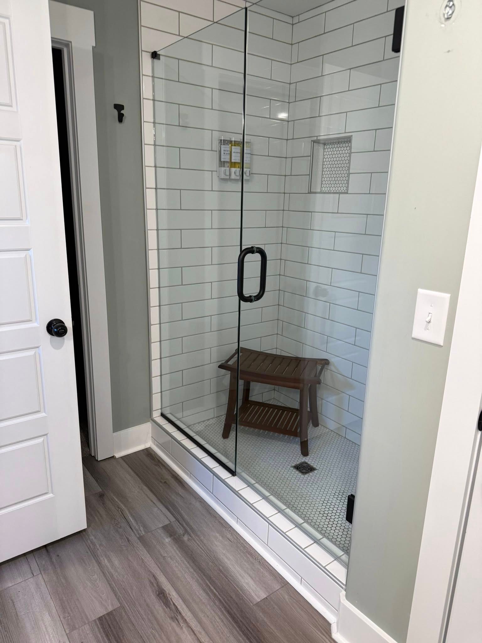 Glass shower enclosure with brick-pattern tile and a wooden stool inside, beside an open bathroom door