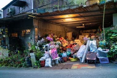 Overcrowded storefront under a awning, piled with bags, boxes, and potted plants at dusk.