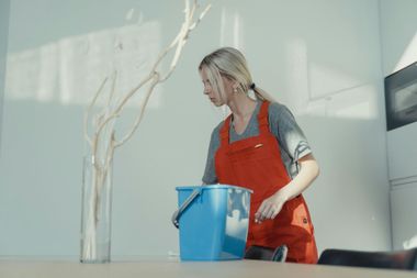Woman in red apron cleaning a bright room with a blue bucket
