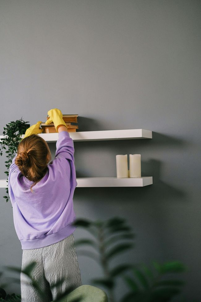 Person in a purple sweater cleaning a white wall shelf with yellow gloves in a gray room