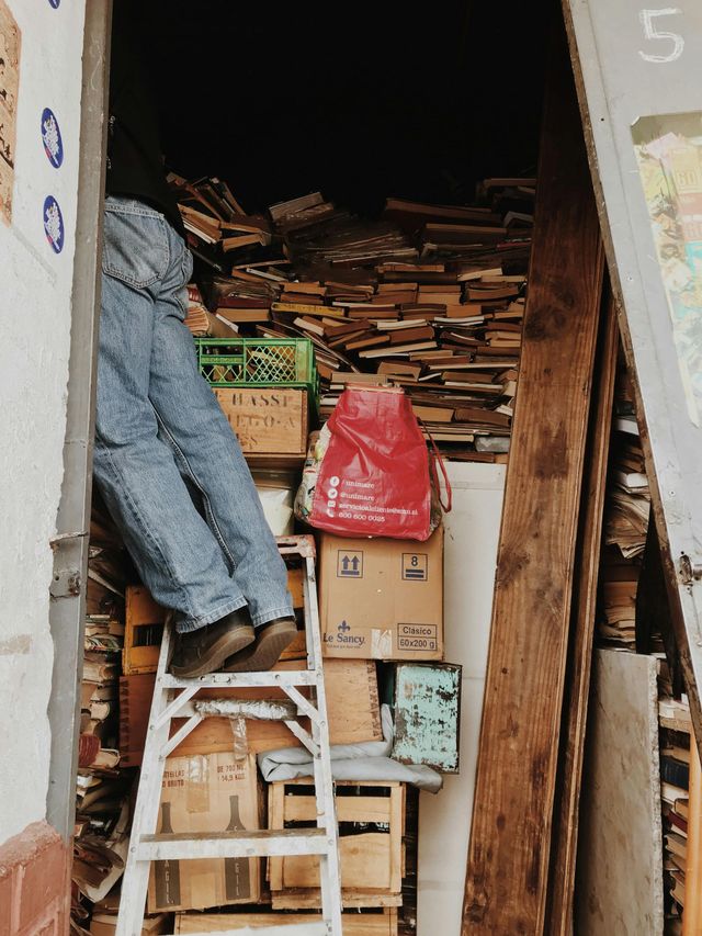 Person on a ladder reaching into a cluttered storage space filled with stacked boxes and wooden planks