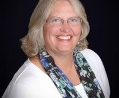 Smiling woman with glasses and blonde hair, wearing a white top and blue patterned scarf against a black background