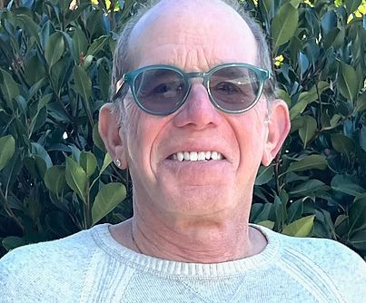 Man wearing sunglasses and a light sweater, smiling in front of leafy greenery