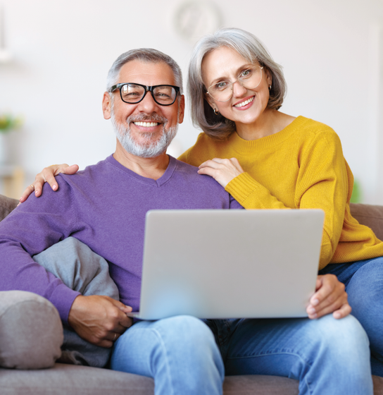 Smiling couple on a couch using a laptop, with the man in a purple sweater and the woman in a yellow sweater.