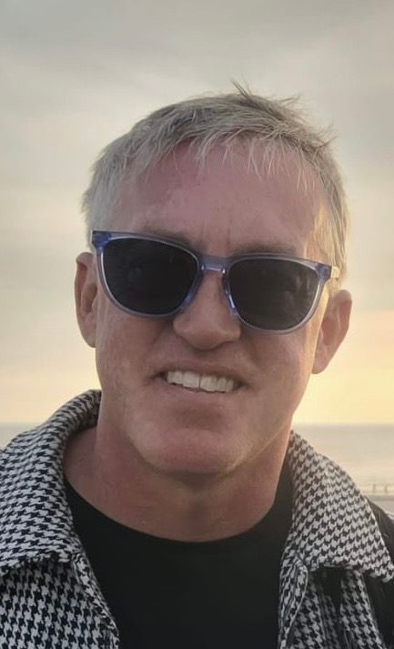 Man wearing sunglasses and a patterned jacket smiles outdoors.