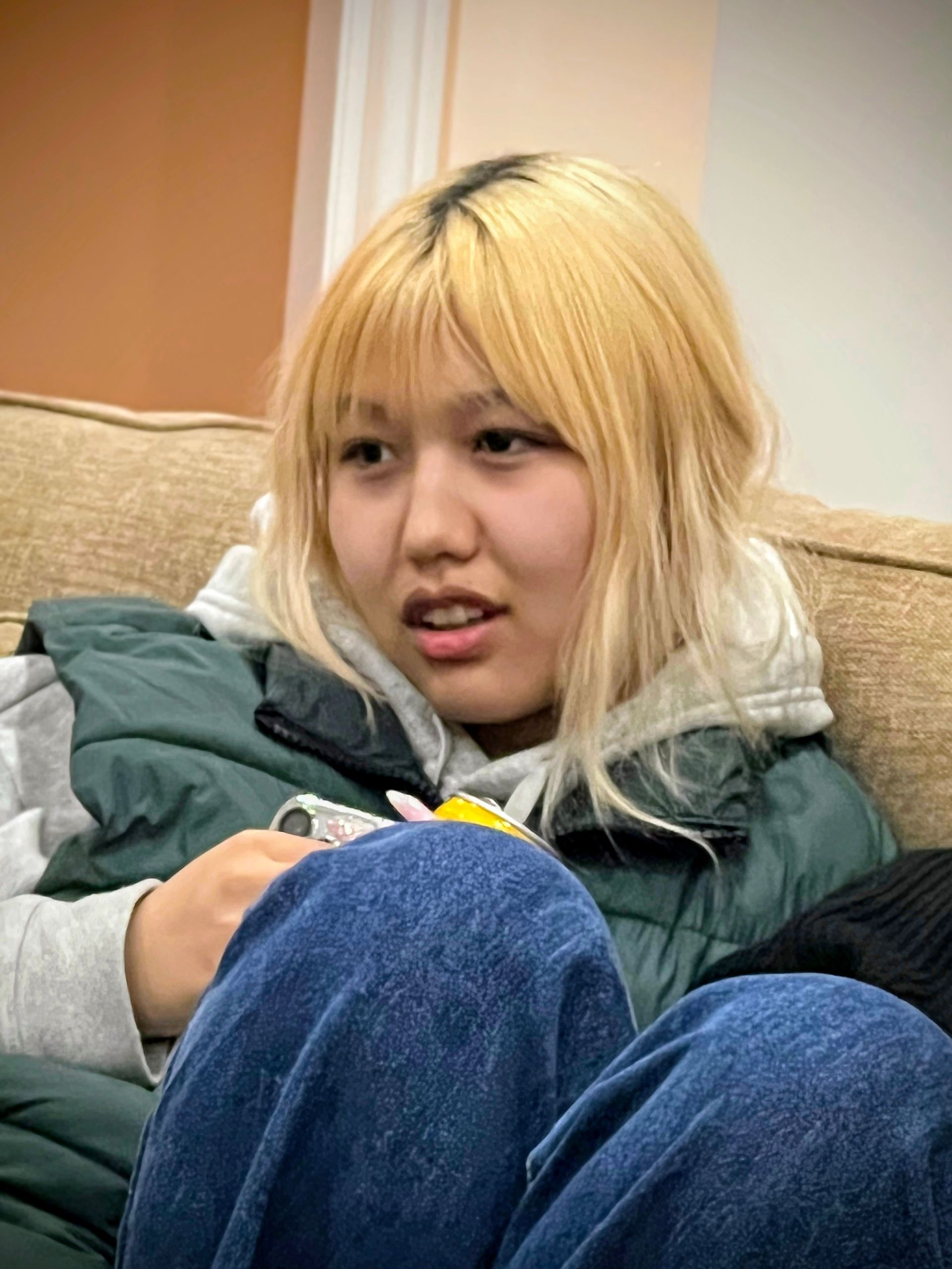 Woman with blonde hair wearing a green vest and jeans, seated indoors, looking slightly concerned.