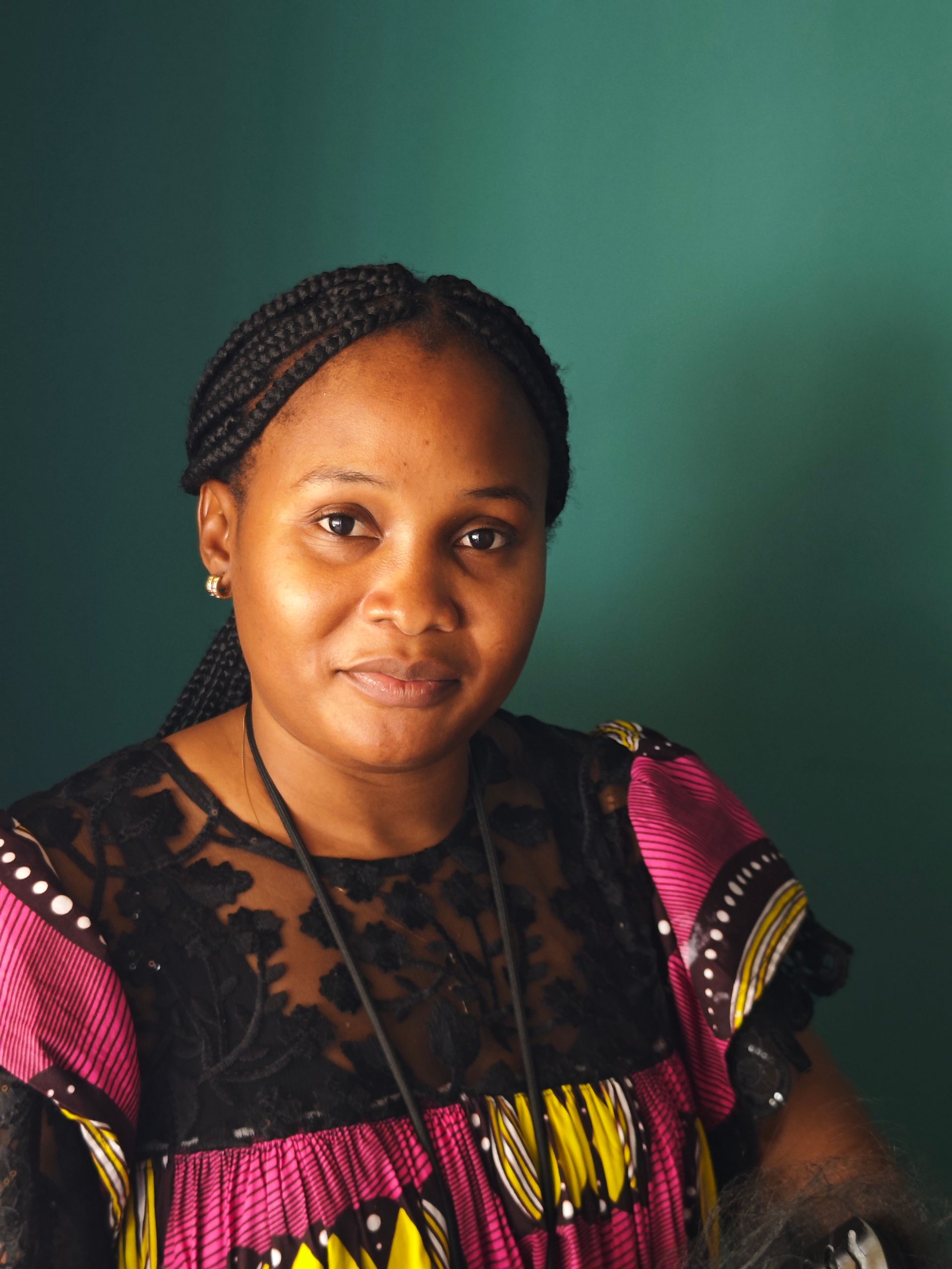 Woman with braided hair, wearing a patterned top with pink and yellow accents, smiles against a green background.