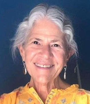 Smiling woman with white hair wearing yellow top and silver earrings.