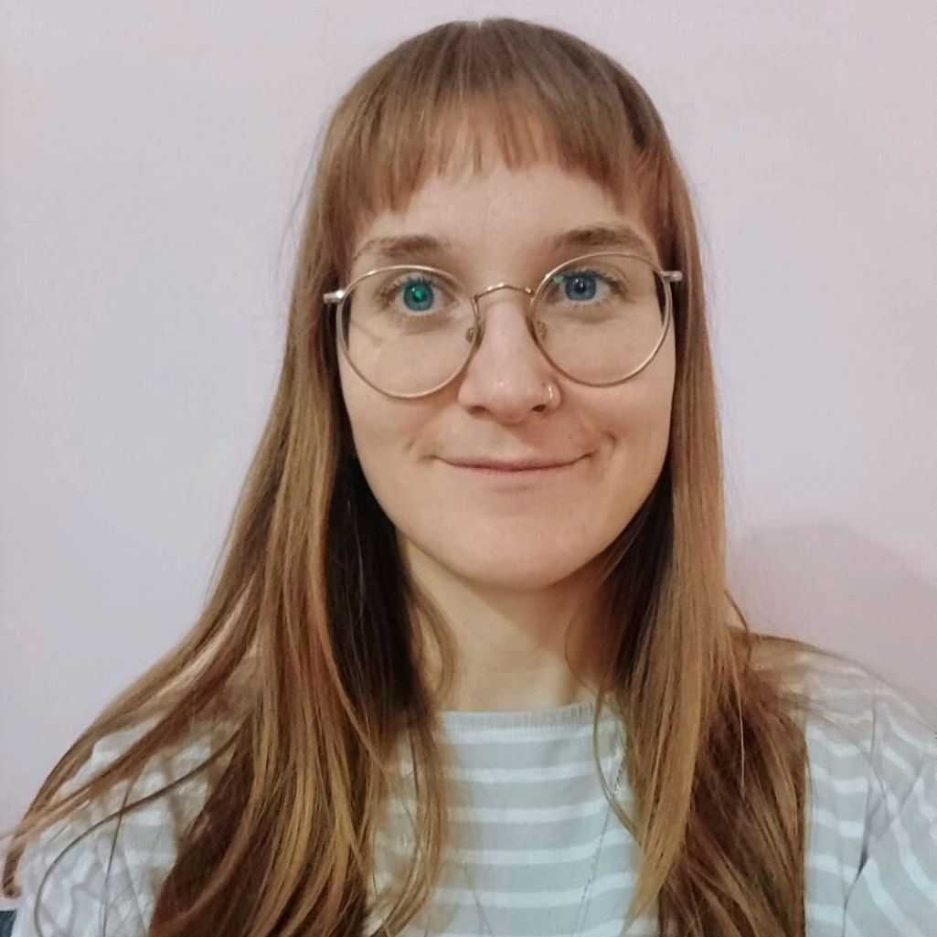 Woman with glasses, bangs, and long brown hair smiles, wearing a striped shirt in front of a pink wall.