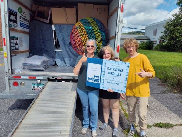 Three people standing by a truck ramp holding a donation sign and colorful woven ball